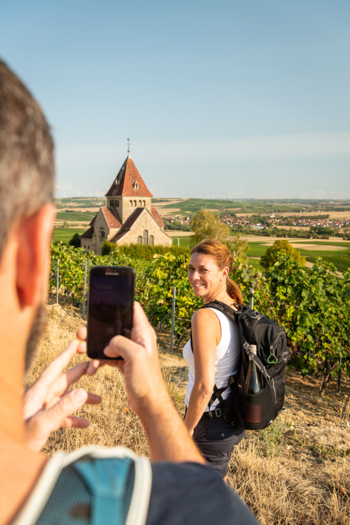 Person photographing a woman with a backpack standing in vineyards near a historic stone church. Rolling fields, rural villages, and rows of grapevines stretch across the landscape under a clear sky.