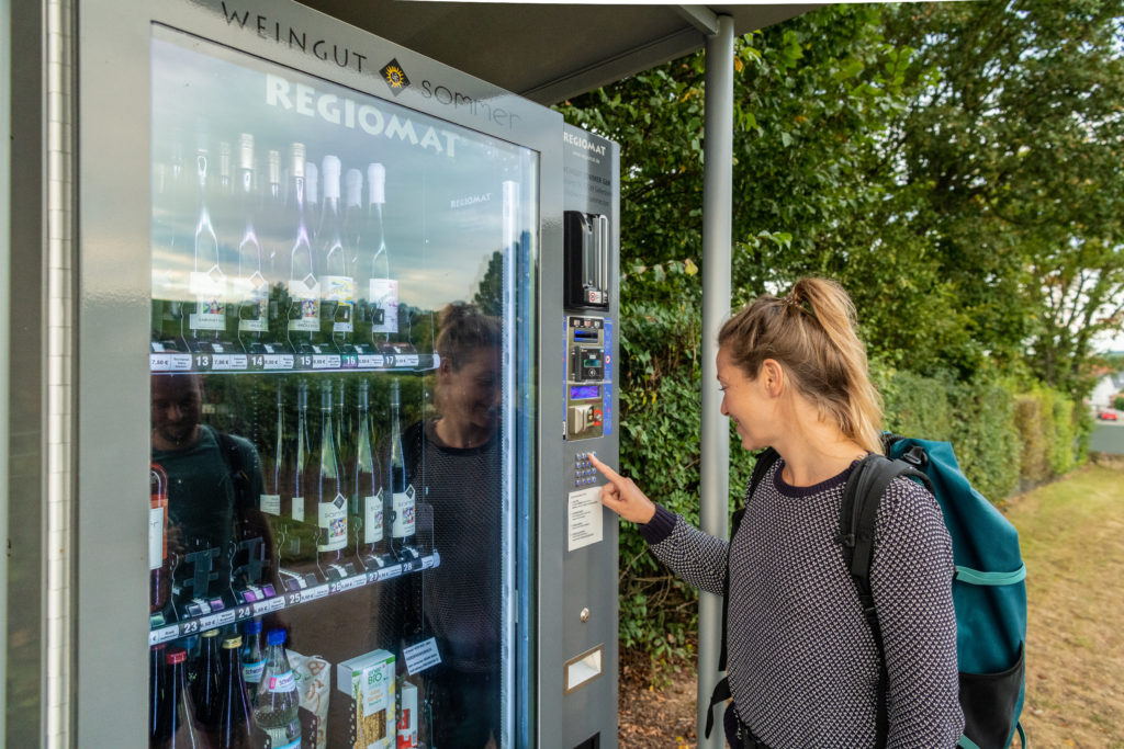 Woman with a backpack using a wine vending machine outdoors beside trees and a roadside path. Bottles of regional wine are visible behind the glass display of the automated self-service machine.