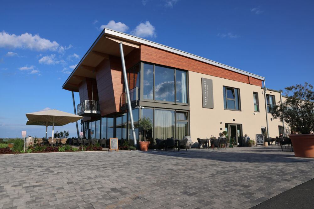 Weinzuhause building with large glass windows, an outdoor terrace, and seating area under a blue sky, surrounded by vineyards and open countryside.