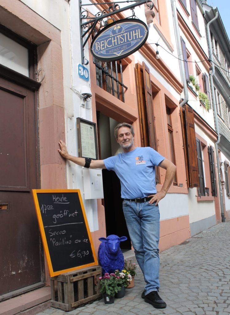 Axel Klug standing outside the Beichtstuhl wine tavern on a cobblestone street, with a chalkboard advertising wine and secco.