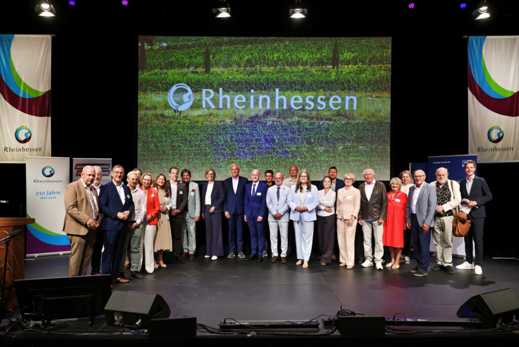 Large group of participants standing on stage at a Rheinhessen conference, with the Rheinhessen logo displayed in the background.